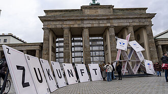 Vor dem Brandenburger Tor steht ein überdimensional großes Kartenhaus, das in sich zusammenbricht
