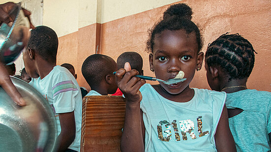 Bild: Ein Mädchen aus Haiti isst eine besonders nahrhafte Mahlzeit und guckt in die Kamera. Hinter ihr sind noch andere Kinder zu erkennen