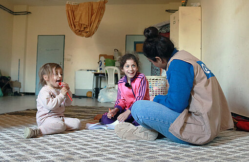Zwei junge Mädchen und eine Mitarbeiterin von Plan International sitzen auf dem Boden und lachen gemeinsam