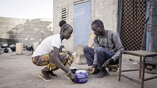 Bandiba und ihr Ehemann sitzen vor ihrem Haus und haben einen Topf vor sich stehen