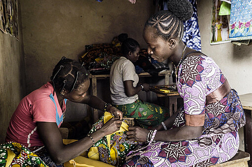 Bandiba sitzt in ihrer Werkstatt vor einer jungen Frau, die etwas mit der Hand näht. Im Hintergrund sitzt eine weitere junge Frau an einer Nähmaschine