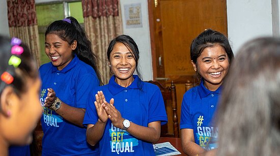 Drei junge Frauen sind im Fokus, sie tragen T-Shirts von Plan International und sind sehr glücklich.