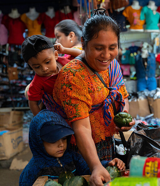 Eine Frau stöbert fröhlich an einem Marktstand. Auf ihren Rücken ist ein kleiner Junge gebunden, neben ihr steht ein weiterer Junge