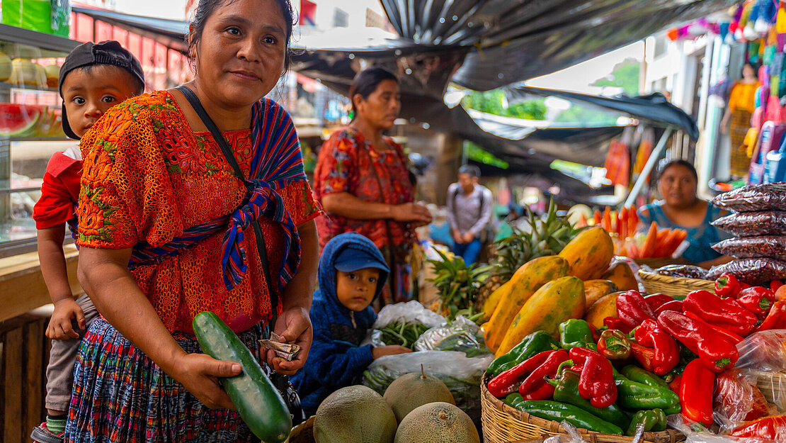 Eine Frau mit zwei kleinen Jungen steht an einem Marktstand mit Obst und Gemüse