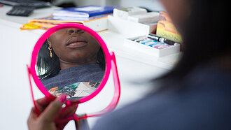 Eine Frau hält einen Kosmetikspiegel in der Hand, man sieht nur einen Teil ihres Gesichts in dem Spiegel